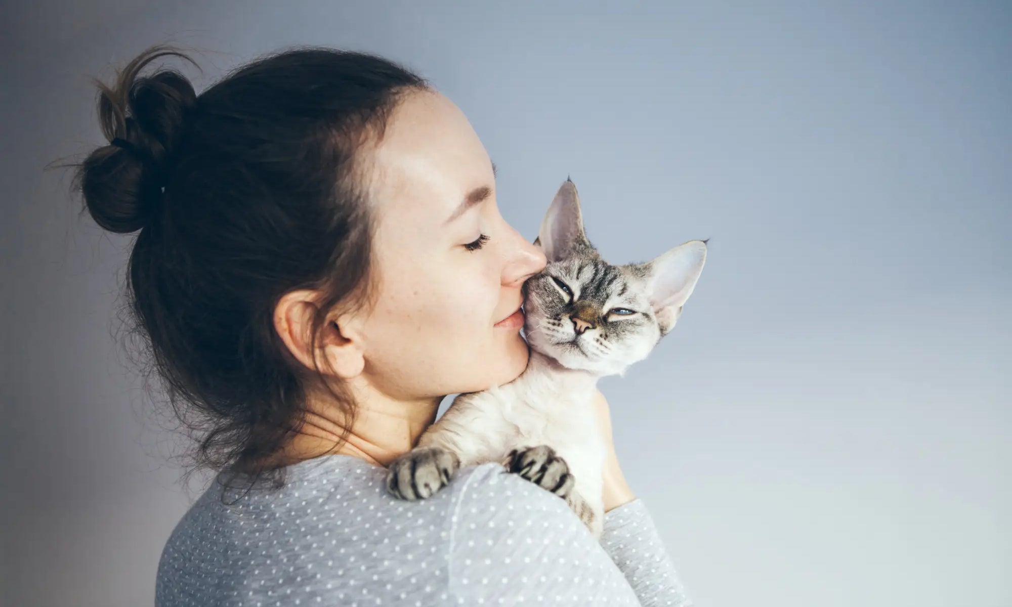 Woman with dark hair kissing a gray and white Devon Rex cat in a cozy setting.