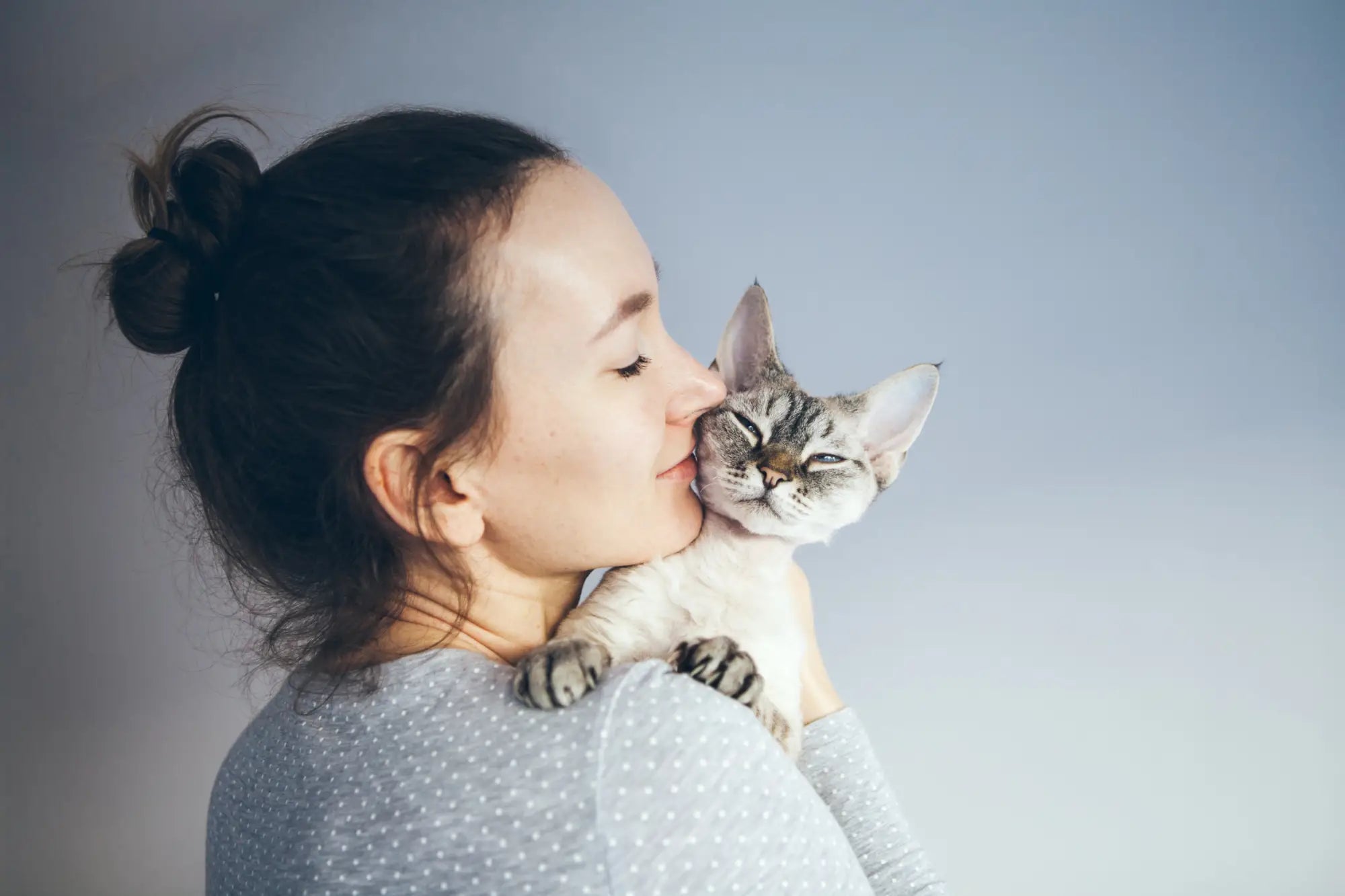 Woman with dark hair kissing a gray and white Devon Rex cat in a cozy setting.