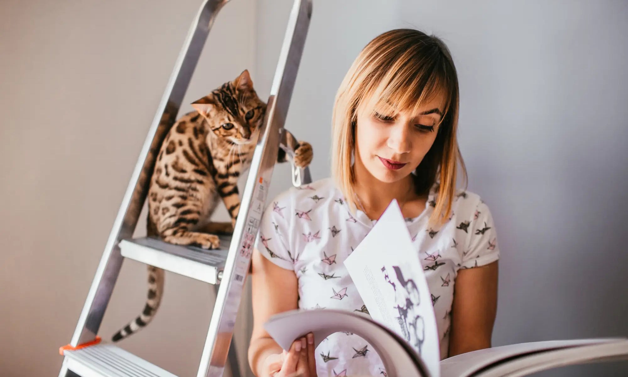 Woman reading a book with a Bengal cat lounging on a ladder beside her.